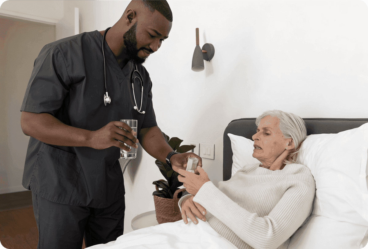 A male nurse in dark scrubs hands a glass of water and medication to an elderly woman in bed.
