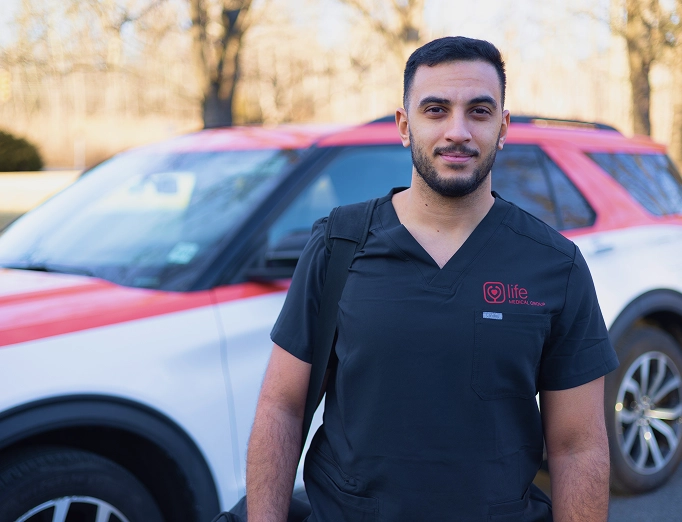 A male paramedic in a red shirt stands next to a medical service car, looking toward the viewer.
