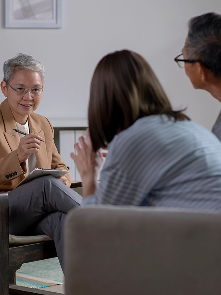 Group of senior adults engaged in a therapeutic session, seated on a couch and armchairs in a bright, comfortable living room setting.