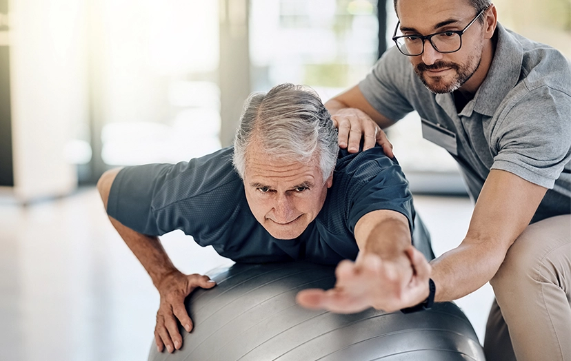 A senior man and his physical therapist doing balance exercises with a large stability ball in a sunlit living room.