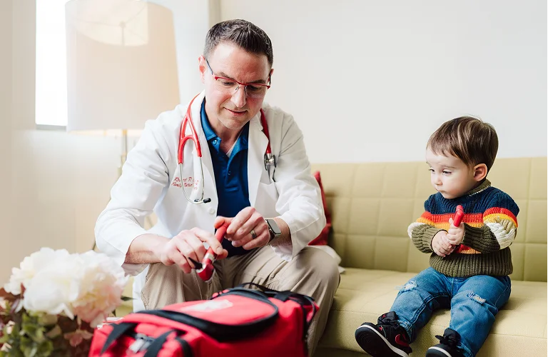 A doctor in a white coat sits on a sofa, looking at a small child's knee, with the child sitting next to him.