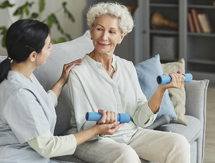 A physical therapist assisting a senior woman with light dumbbell exercises on a couch in her home