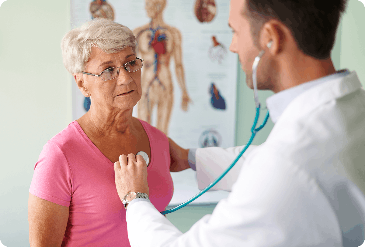 A male doctor in a white coat listening to an elderly woman's heart with a stethoscope, with an anatomical chart of the human circulatory system visible in the background.